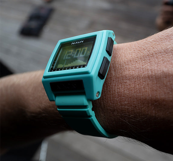 A person's arm wears a turquoise Nixon digital watch with a black face showing 12:00.