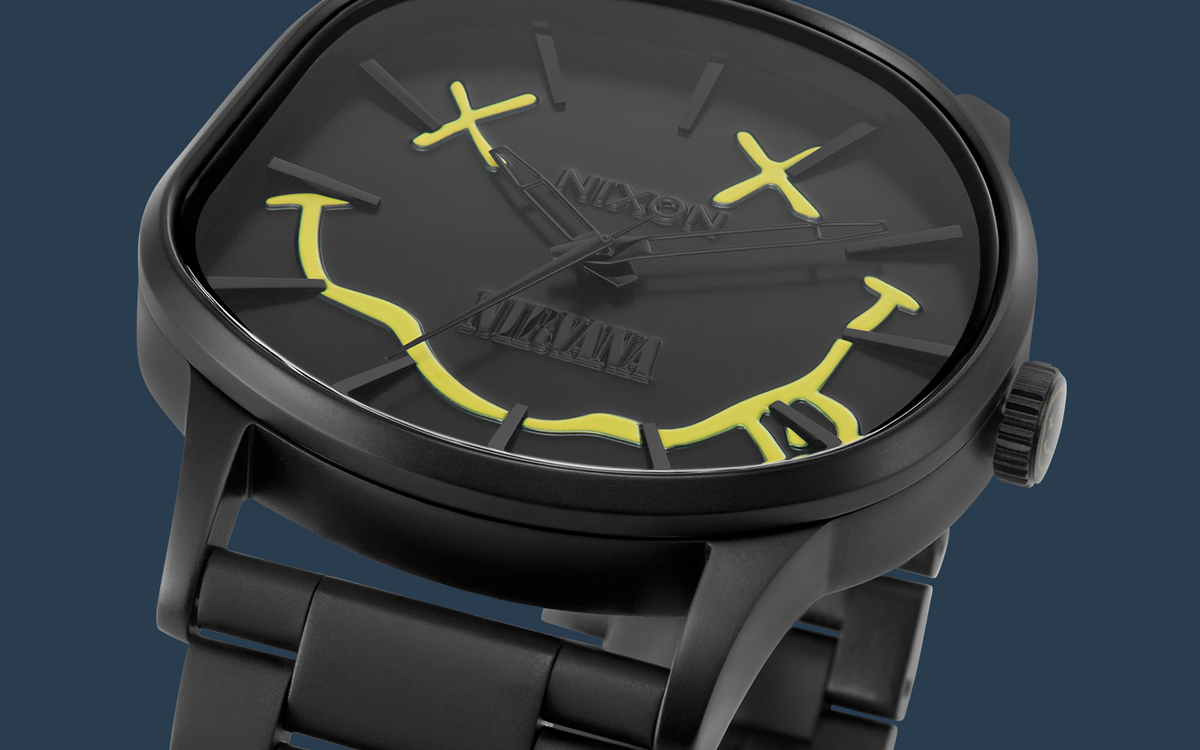 A close-up of a Nixon X Nirvana watch with a matte black case and link bracelet, featuring a black dial with bright yellow graphic accents and a day-date window.