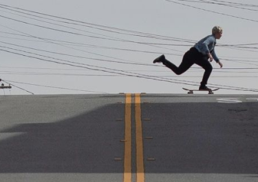 A person in a light blue shirt and dark pants rides a skateboard up a steep street with parked cars on both sides and utility poles in the background.
