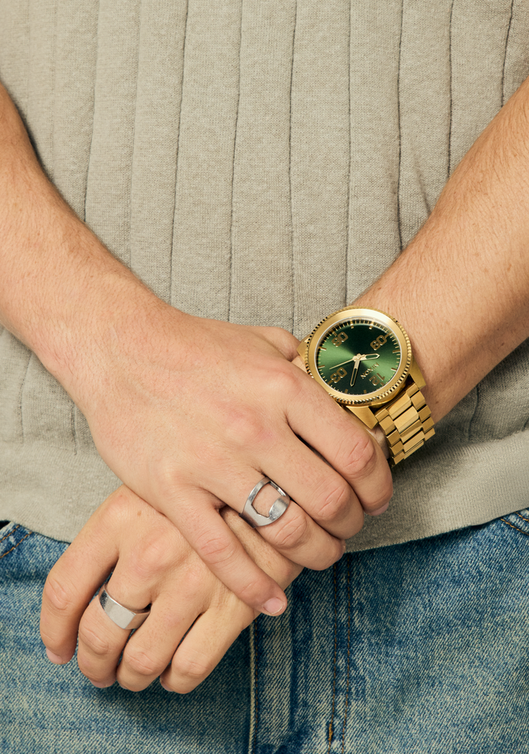 Man has hands folded in front of him while wearing a gold and green Nixon Corporal Stainless Steel analog watch