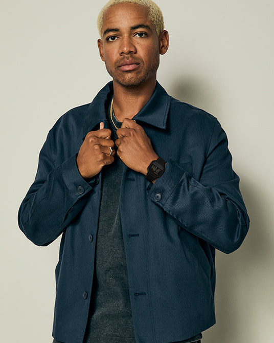 Man wearing a dark blue jacket with Black Nixon watch against a plain background