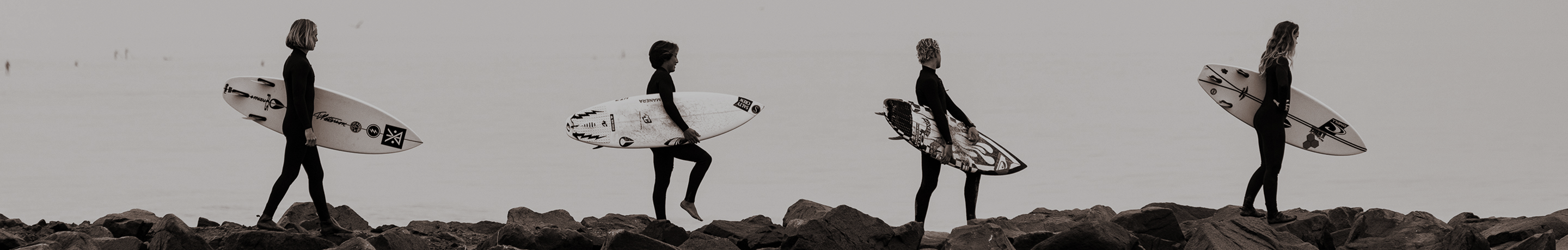 Four surfboarders walk along the ocean while holding their surfboards.